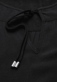 Zwarte blouse met een gestructureerd oppervlak, voorzien van een V-hals en een trekkoord met metalen accenten. Gladde stof, subtiel patroon.
