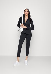Zwarte op maat gemaakte blazer met revers, gecombineerd met bijpassende strakke broek. Een witte schoudertas en witte sneakers maken de look compleet.