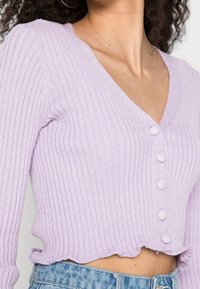 Donna che indossa un cardigan a V rivestito in lavanda con bottoni rivestiti in tessuto e orlo arricciato, abbinato a jeans blu chiaro.