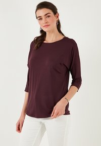 Burgundi hosszú ujjú felső, puha, könnyű anyagból, lekerekített nyakkivágással és háromnegyedes ujjakkal, fehér nadrággal párosítva.