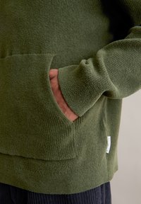 Πράσινο φούτερ με πλέξη, με υφή στην επιφάνεια, διαθέτει μπροστινή τσέπη και ριμπ μανίκια. Ο ιστός φαίνεται μαλακός και ζεστός.