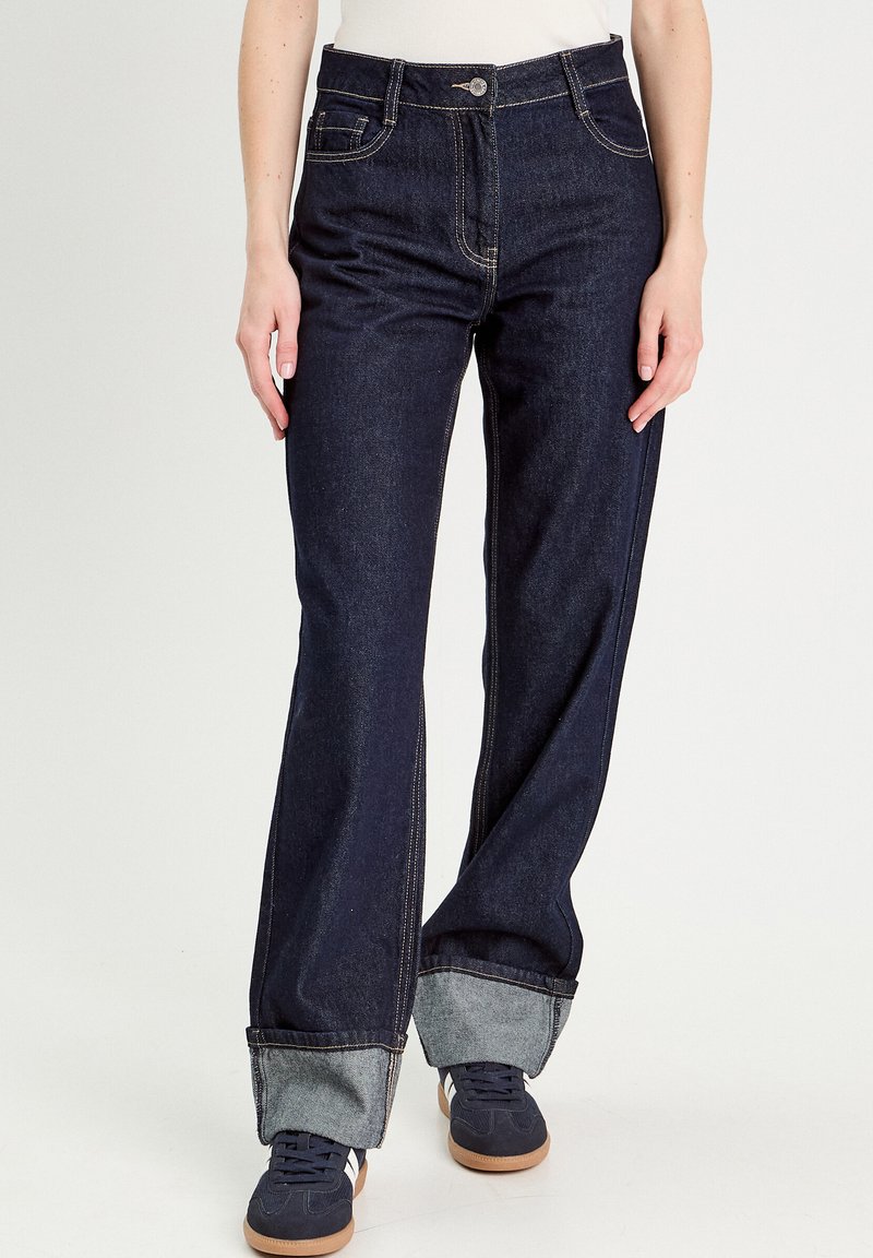Dunkelblaue weit geschnittene Jeans mit einem hochgerollten hellblauen Saum. Verfügt über einen Reißverschluss, einen Knopfverschluss und fünf Taschen. Hergestellt aus Denim-Stoff.