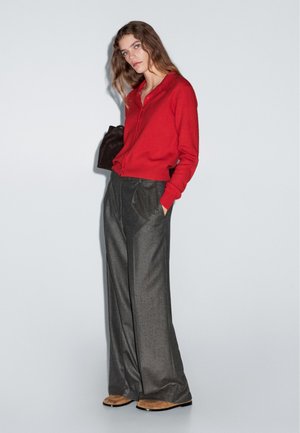 Cardigan rosso in lana con dettagli di bottoni, abbinato a pantaloni grigi a vita alta. Lo stile include gambe ampie e una texture sottile. Le scarpe color cuoio completano il look.