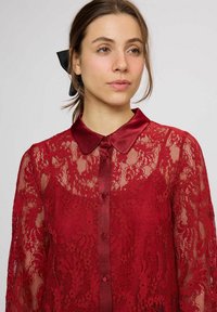 Bluză roșie din dantelă, cu guler din satin și nasturi, mâneci lungi și modele florale sofisticate pe toată suprafața țesăturii.