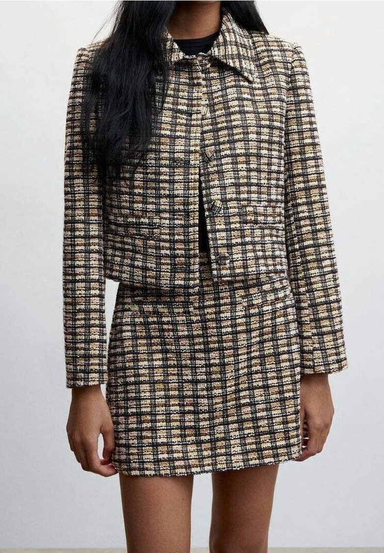 Femme portant une veste courte à carreaux beige, noire et blanche avec une jupe assortie, debout devant un fond clair uni.