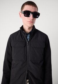 Jachetă neagră cu fermoar, dotată cu două buzunare pe piept și o textură netedă, purtată cu ochelari de soare negri, mari și unii unghiulari, și o cămașă albastră dedesubt.