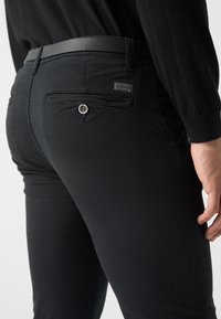 Pantaloni neri a sigaretta in cotone con una texture liscia, dotati di una tasca posteriore con bottone e una vita accentuata in pelle.
