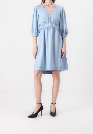 Mujer vestida con un vestido azul claro hasta la rodilla, con mangas abullonadas hasta el codo y zapatos de tacón negro con pulsera en el tobillo.