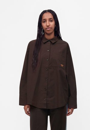 Personne aux longs cheveux tressés portant une chemise marron foncé oversize boutonnée avec un petit logo sur la poche poitrine.