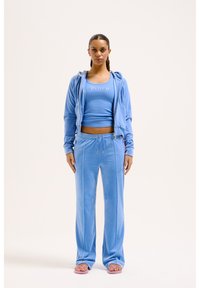 Velours trainingspak in lichtblauw, met een rits-hoodie en een aansluitend tanktop met "Juicy" tekst, gecombineerd met bijpassende broeken en roze sandalen.