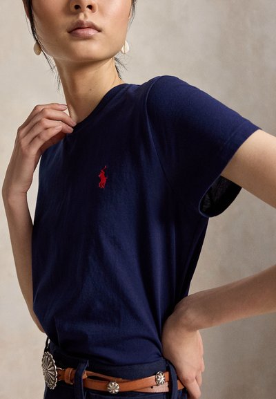 Γυναίκα που φοράει μπλε σκούρο T-shirt με κόκκινο κεντημένο λογότυπο, συνδυασμένο με καφέ δερμάτινη ζώνη με ασημένια conchos, πόζαρε με το χέρι κοντά στον λαιμό.