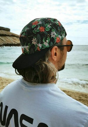 Schwarze Kappe mit buntem Blumenmuster, das rote und weiße Blumen zeigt, kombiniert mit einem weißen Hemd und Sonnenbrille, vor einem Strandhintergrund.