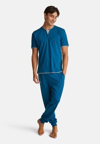 Pánská dvoudílná sada na odpočinek v tónu teal, která zahrnuje tričko s krátkým rukávem a knoflíkovým límcem ve stylu henley a kalhoty na šňůrku. Měkká látka, volný střih.