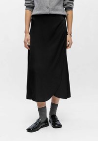 Sort midi-nederdel med wrap-design, glat stof og en let glans. Kombinationen med sorte sko og grå sokker, båret med en grå sweater.