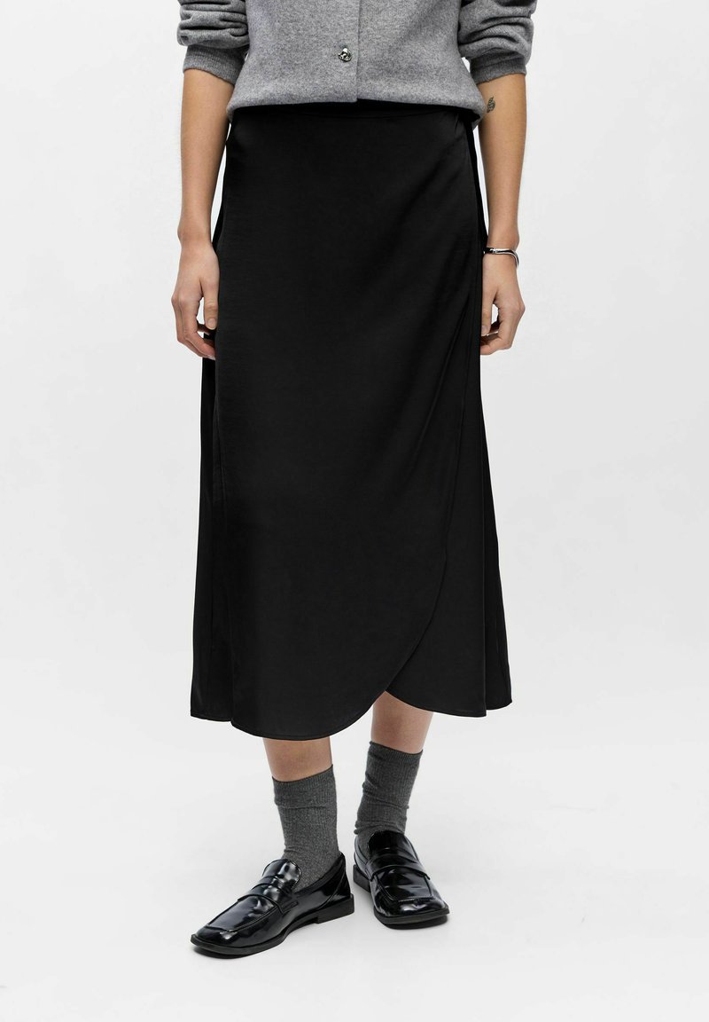Sort midi-nederdel med wrap-design, glat stof og en let glans. Kombinationen med sorte sko og grå sokker, båret med en grå sweater.