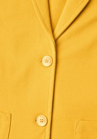 Blazer jaune avec deux boutons étiquetés "Street One" et deux poches avant visibles près des bords inférieurs de l’image.
