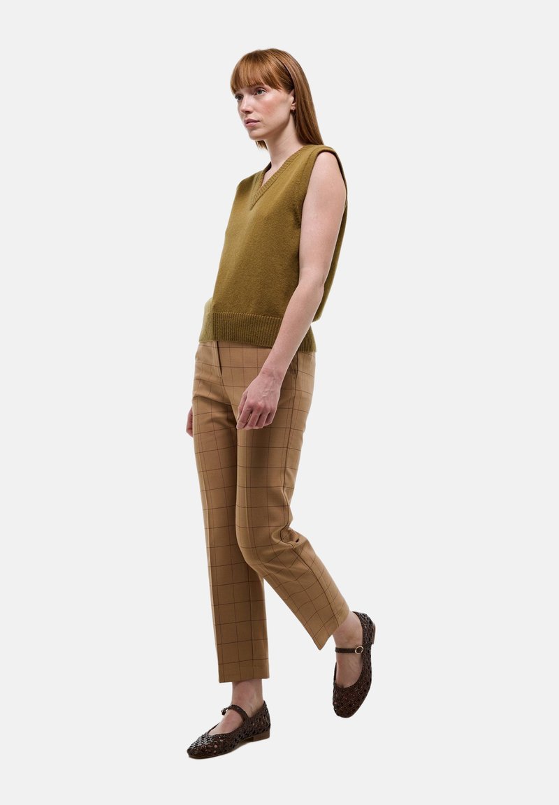 Un pull sans manches vert olive avec un col en V, associé à un pantalon à carreaux beige. Des ballerines tressées marron avec une sangle complètent le look.