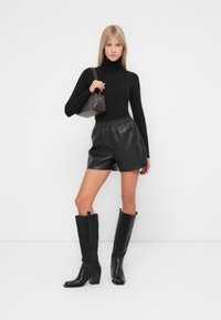 Zwarte coltrui, zwarte neplederen shorts met elastische tailleband, kniehoge zwarte haklaarzen en een kleine bruine handtas.