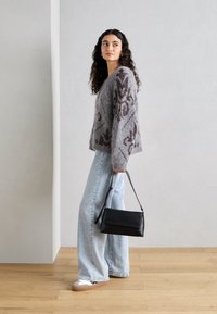 Grauer Strickpullover mit braunem Muster, hellblaue weitgeschnittene Jeans und schwarze Handtasche. Das Model steht vor einem neutralen Hintergrund.