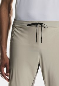 Pantaloni beige chiaro realizzati in tessuto liscio con chiusura a cordoncino, caratterizzati da dettagli neri. Primo piano della vita e posizione della mano.