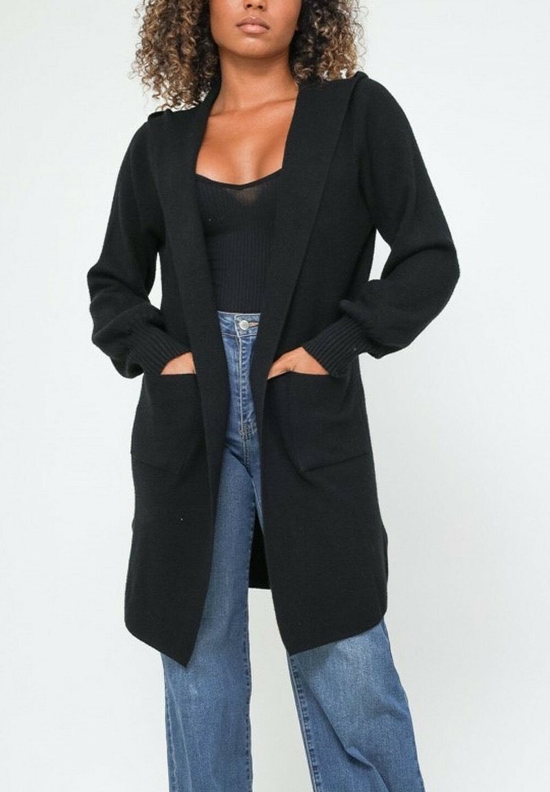 Femme debout portant un long cardigan noir avec poches sur un haut noir et un jean bleu, les mains posées à l'intérieur des poches du cardigan.