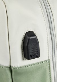 Tela suave de color gris claro con un panel verde. Presenta un puerto USB negro con líneas grabadas y un borde cosido. Detalle de cremallera visible.