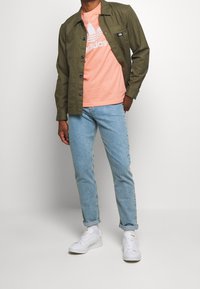 Olivgrön skjortjacka över en persikofärgad t-shirt med vit logotyp, kombinerad med ljusblå jeans och vita sneakers. Avslappnad outfit.