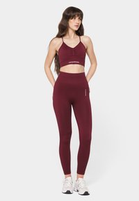 Burgundy sports bra with gathered front and logo, paired with high-waisted leggings. Smooth texture, fitted design, and contrasting white accents.