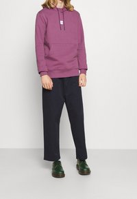 Lila huvtröja med en framficka, matchad med marinblå vida byxor och gröna skor. Huvtröjan har en liten logodetalj.