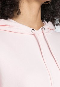 Primo piano di collo e spalle con capelli ricci, che indossa un hoodie rosa con cordini e un orecchino a cerchio dorato.