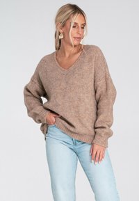 Kobieta z blond włosami ma na sobie luźny brązowy sweter z dzianiny, jasnoniebieskie dżinsy, złote kolczyki w kształcie kół oraz naszyjnik, pozując z jedną ręką w kieszeni.