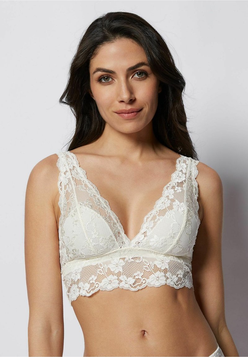 Bralette en dentelle de couleur blanc cassé, avec un décolleté V profond et de larges bretelles en dentelle. Des motifs floraux ornent la dentelle et l'ourlet, ajoutant texture et détail.