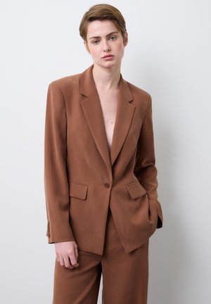 Jonge persoon met kort haar, gekleed in een bruine colbert en bijpassende broek, staand tegen een effen witte achtergrond.