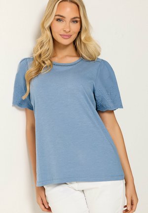 Blonde Frau trägt ein hellblaues kurzärmeliges Oberteil mit floralen Stickärmeln und weiße Hose, steht vor einem einfarbigen Hintergrund.