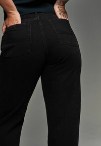 Zwarte denim broek met een recht gesneden ontwerp, voorzien van achterzakken en subtiele stikdetails. De stof heeft een gladde textuur.