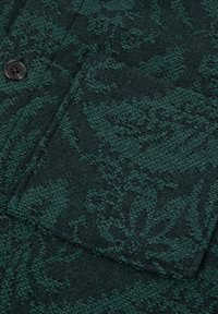Tessuto verde scuro con texture e motivo floreale intessuto, con bottone e contorno della tasca.
