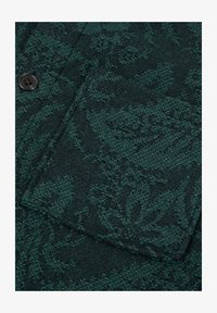 Tejido texturizado de color verde oscuro con un patrón floral tejido, que presenta un botón y un contorno de bolsillo.