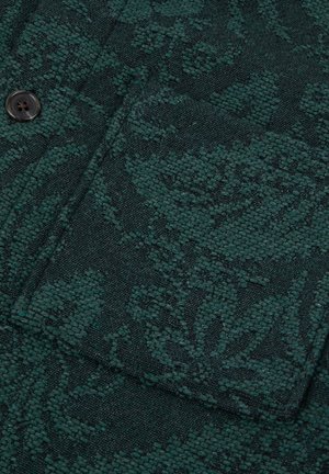 Donkergroen textiel met textuur en een geweven bloemenpatroon, met een knoop en een zakomtrek.
