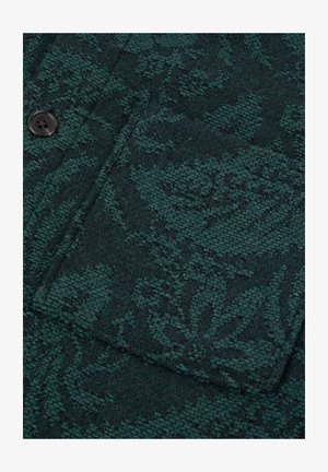 Donkergroen textiel met textuur en een geweven bloemenpatroon, met een knoop en een zakomtrek.