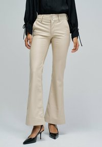 Femme portant un pantalon beige taille haute évasé, un haut noir à manches longues et des escarpins noirs à bout pointu se tenant contre un fond uni.