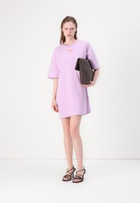 Lilla oversized t-skjortekjole med korte ermer og en liten oransje logo. Modellen holder en brun geometrisk clutch og går med svarte sandaler med stropper.