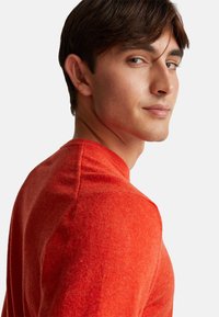 Młody mężczyzna z krótkimi brązowymi włosami, ubrany w pomarańczowy sweter, lekko odwraca głowę, patrząc na kamerę na białym tle.