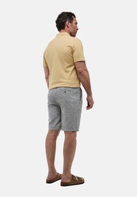 Un polo beige clair, associé à un short rayé gris clair. Sandales marron à plusieurs brides. L'homme est de dos, tourné vers la gauche.
