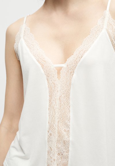 Femme portant un haut blanc sans manches avec un profond décolleté en V bordé de délicats détails en dentelle.