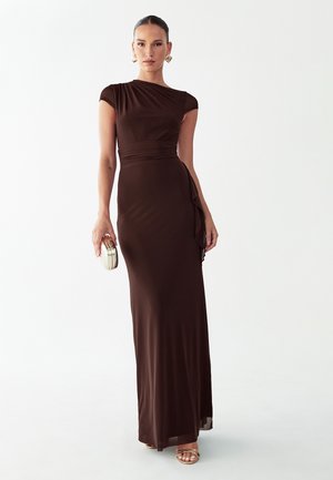 Mujer de pie con un vestido ajustado de largo hasta el suelo de color marrón oscuro, sosteniendo un pequeño bolso de mano dorado, y llevando sandalias y pendientes dorados.