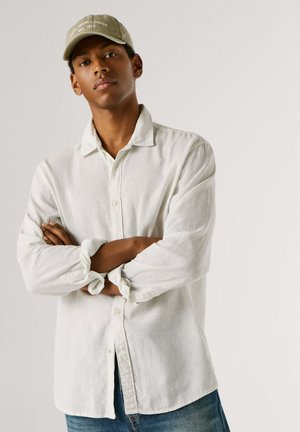 Jauns vīrietis ar saliktām rokām, valkājot gaišas krāsas pogājamu kreklu, zaļu cepuri un zilas džinsas, vienkārša fona priekšā.