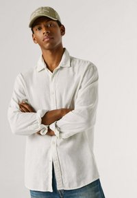 Jaunuolis sukryžiavęs rankas, dėvintis šviesios spalvos marškinius, žalią kepurę ir mėlynas džinsus, stovintis priešais vienodą foną.