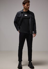 Svart flanellskjorta över en svart t-shirt, svarta byxor och svarta snörskor. Klädseln har en minimal etikett på skjortan.