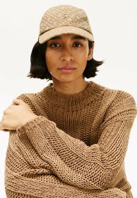 Maglione marrone lavorato a maglia con design testurizzato, indossato con un cappello beige decorato con un motivo e un logo sul davanti. Casual, vestibilità rilassata.
