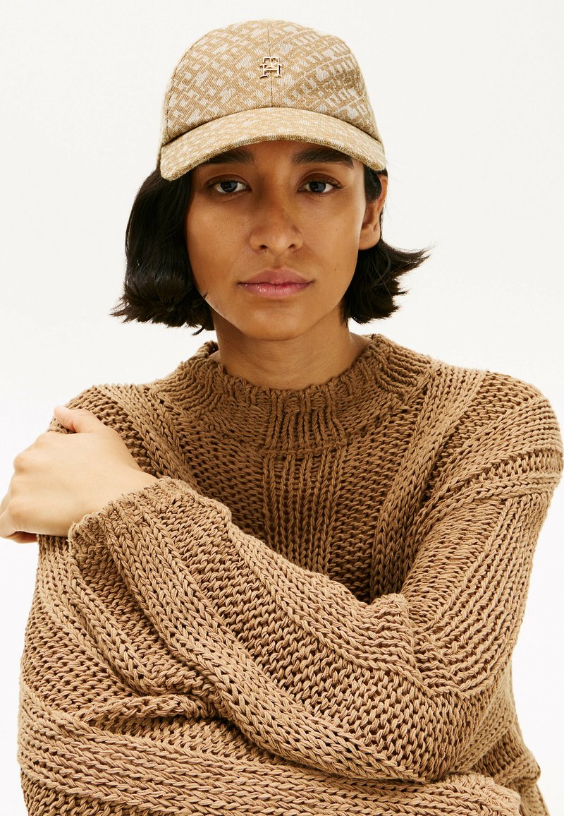 Brauner, gestrickter Pullover mit strukturiertem Design, getragen mit einer beigen, gemusterten Mütze, die ein Logo auf der Vorderseite hat. Lässiger, lockerer Schnitt.